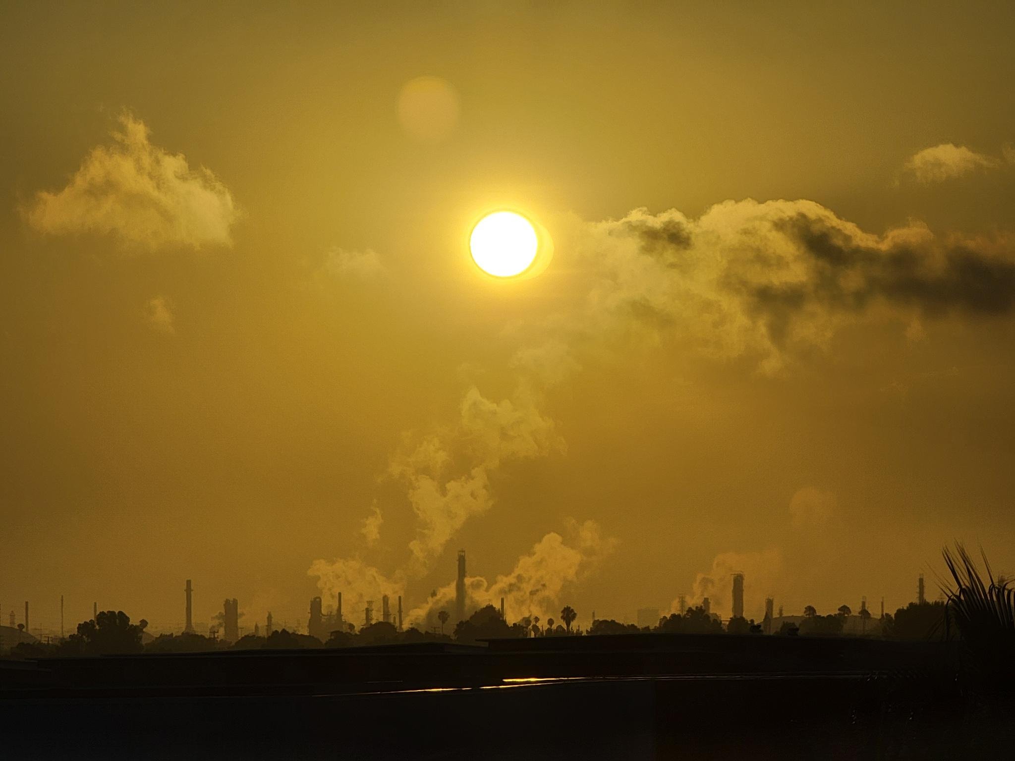 An oil refinery against a yellow sky and sun in the morning.
