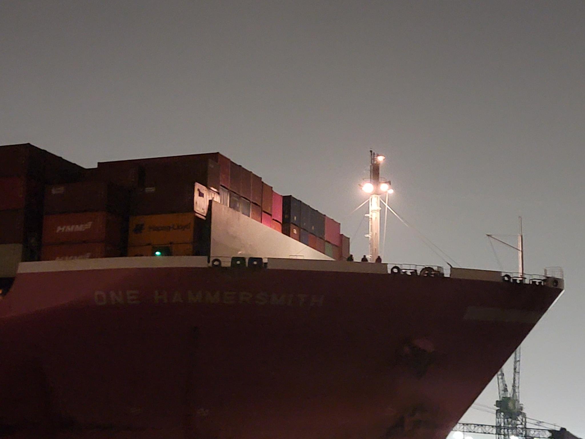 The bow of a container ship at night, illuminated by deck lights.